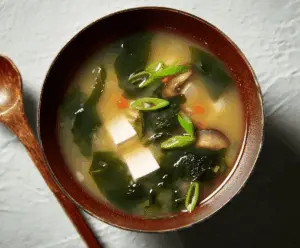 A bowl of steaming miso soup garnished with sliced green onions and tofu cubes, served in a traditional ceramic bowl on a wooden table.